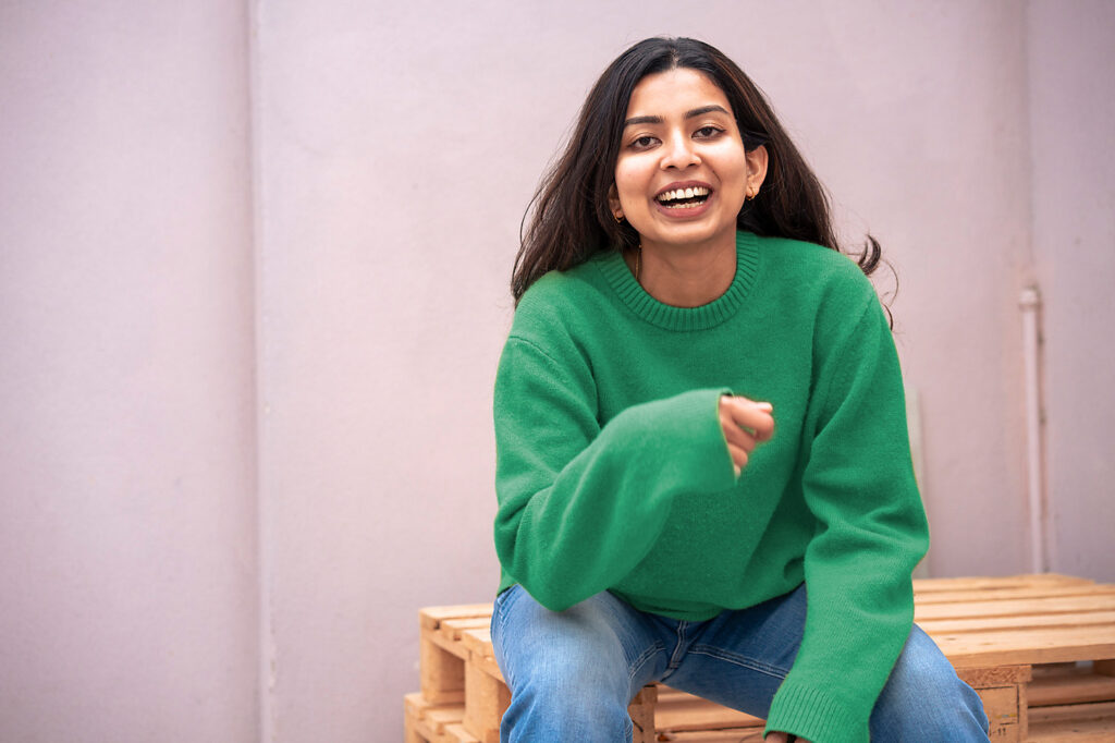 junge Frau mit Jeans und grünem Pullover sitzt lachend auf einem Stapel Paletten