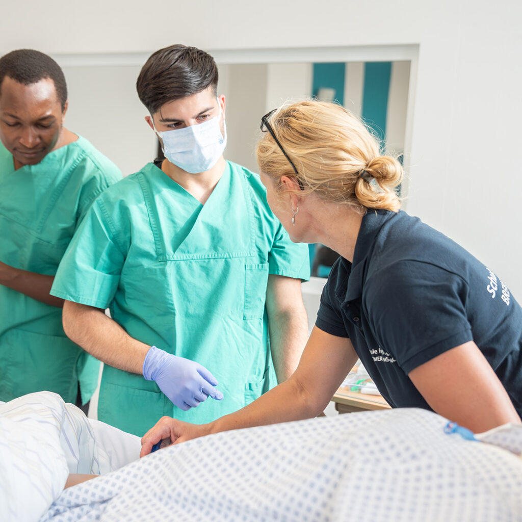 Praxisanleiterin mit angehenden Pflegefachmännern an einem Pflegebett
