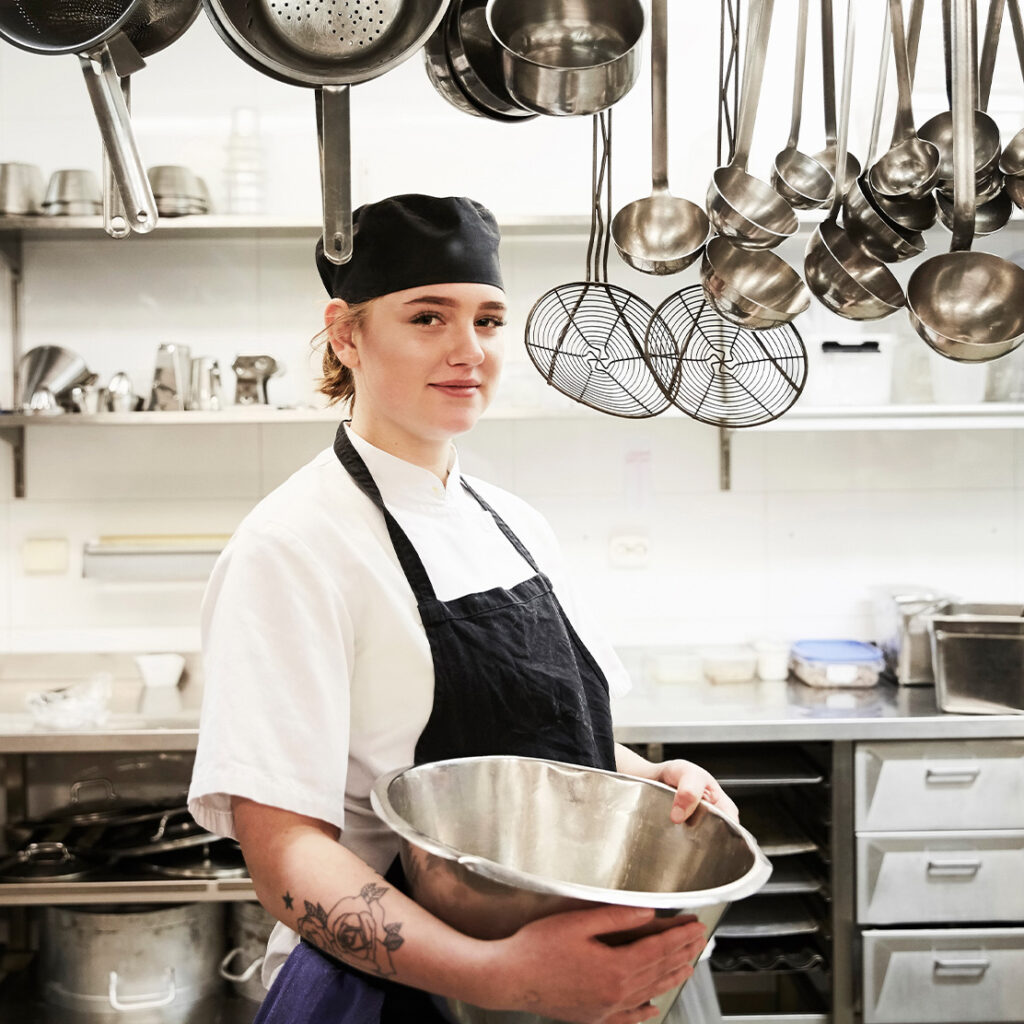 junge Frau in Berufskleidung, mit einem großen Gefäß in der Hand, in einer Gastronomieküche
