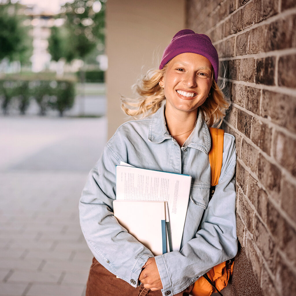 junge Frau mit blonden Haaren lehnt lächelnd an einer Wand